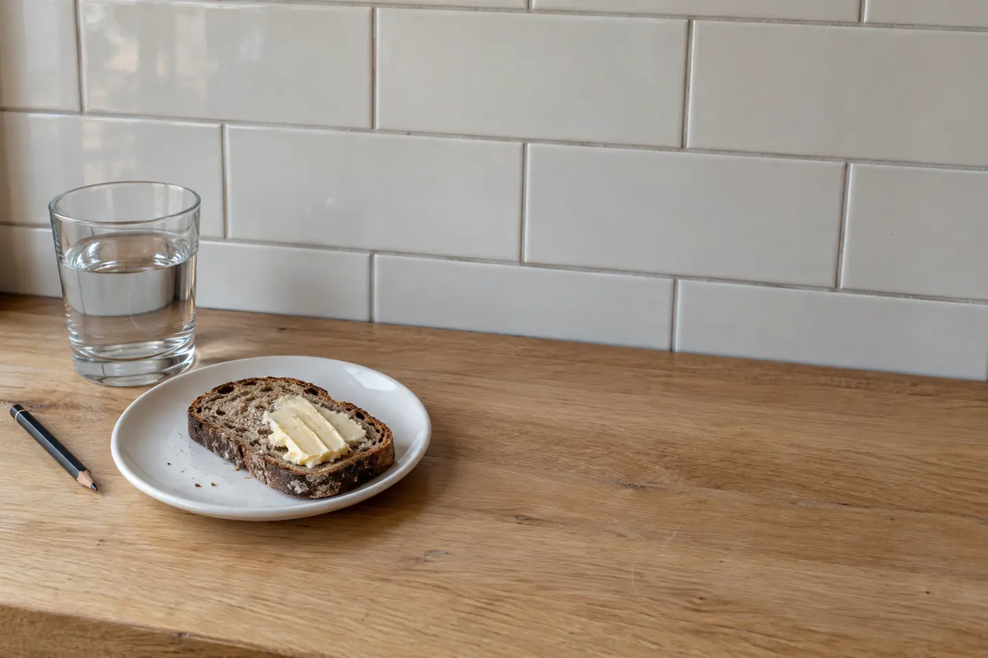 A quiet bistro counter with one rye and butter plate, a water tumbler and a pencil.